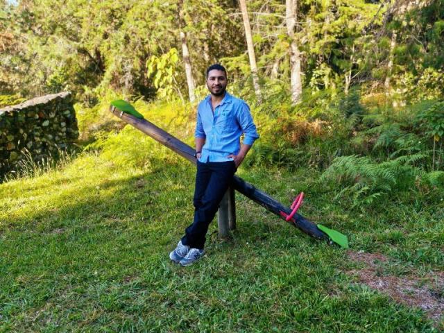 Edahi está de pie con una sierra de corte verde y marrón apoyada contra un poste de madera, vistiendo una camisa azul, pantalones negros y zapatillas grises, en un bosque cubierto de hierba..