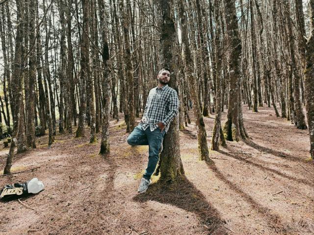 Edahi lehnt an einem Baum in einem Wald, trägt ein kariertes Hemd, Jeans und Turnschuhe, hat ein Bein angehoben und wirkt entspannt und lässig, mit einem leichten Bart..