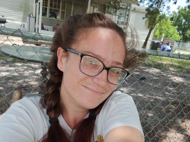 SpicyRedHead has a smug smile, brown braided hair, glasses, and a white shirt. She's in front of a chain-link fence with a house in the background.