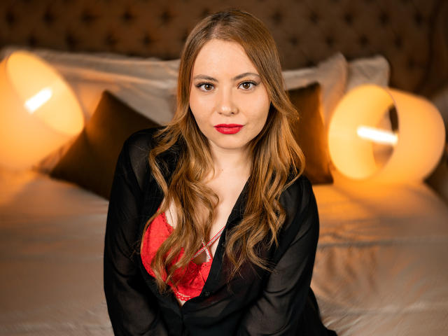 KatteAdams con reggiseno di pizzo rosso, vestaglia trasparente nera e rossetto rosso, seduta sul letto con lampade arancioni calde, lunghi capelli biondi ondulati e sguardo seducente..
