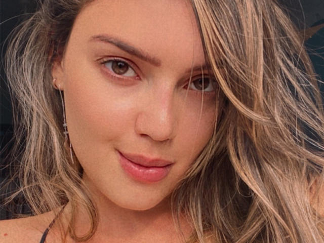 Miabem's face, blonde wavy hair, light skin, brown eyes, slight smile, pink lips, small hoop earrings, soft makeup, visible nose, and gentle expression.