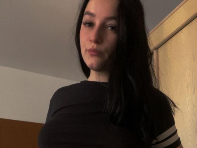 LeahBrooks with black hair, wearing a black shirt with white stripes, has a cute face and subtle smile, small nose, and light skin, standing in a dimly lit room.