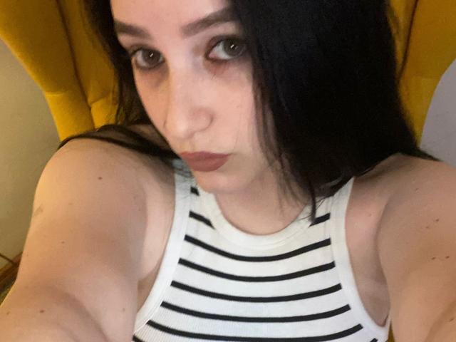 LeahBrooks with dark hair, pouty lips, wearing a white striped tank top, arms raised, sitting in a yellow chair, looking up at the camera. Her tits are partially visible.
