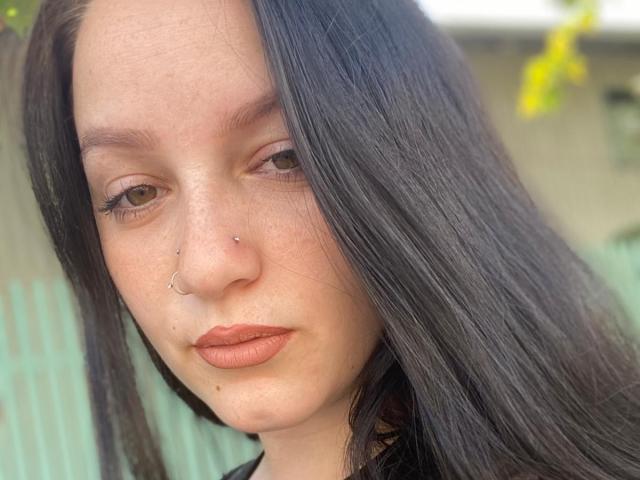 LeahBrooks has long black hair, brown eyes, freckles, a nose ring, and a silver nostril piercing. She's wearing brown lipstick and looking directly at the camera.