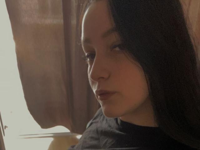 LeahBrooks with dark hair, light skin, and pouty lips, wearing a black shirt, gazing slightly to the side, soft light from the left, brown curtain in the background.