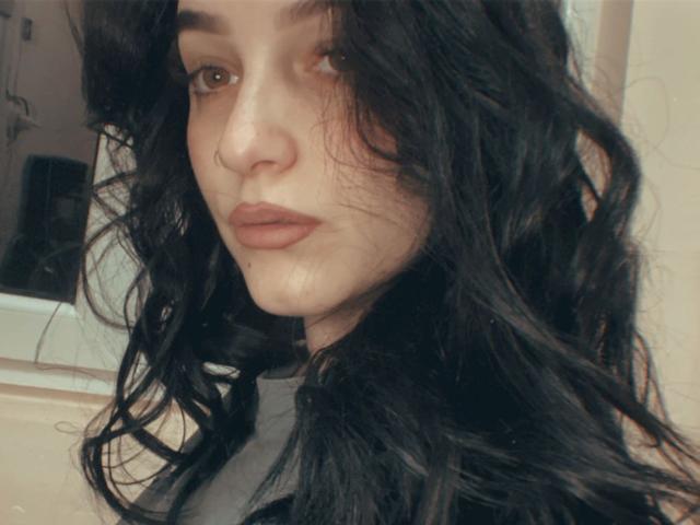 LeahBrooks has wavy black hair, a nose ring, and brown eyes with subtle makeup and pouty lips. Her skin is fair, and she's looking to the side.