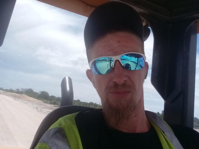 SpicyRedHead, wearing reflective sunglasses, black cap, and neon green vest, has a light beard and is sitting in a vehicle with a cloudy sky outside.