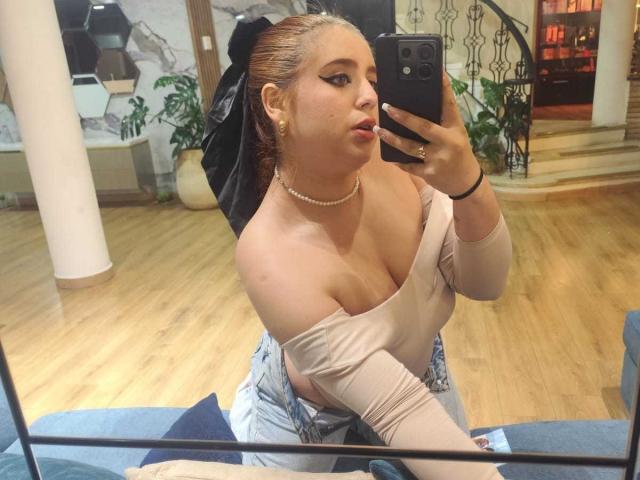 StephanieMoretti with light skin, large tits, and a black bow in her blonde hair, wearing a beige off-shoulder top, taking a mirror selfie with a pearl necklace.