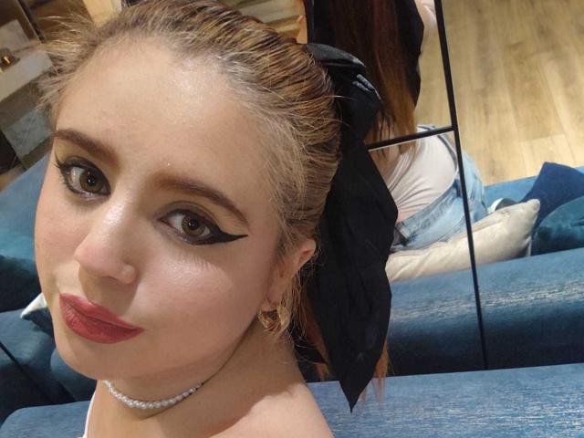 StephanieMoretti has light brown hair in a black ribbon, thick black eyeliner, red lipstick, and a pearl necklace, sitting on a blue couch with wooden floors.