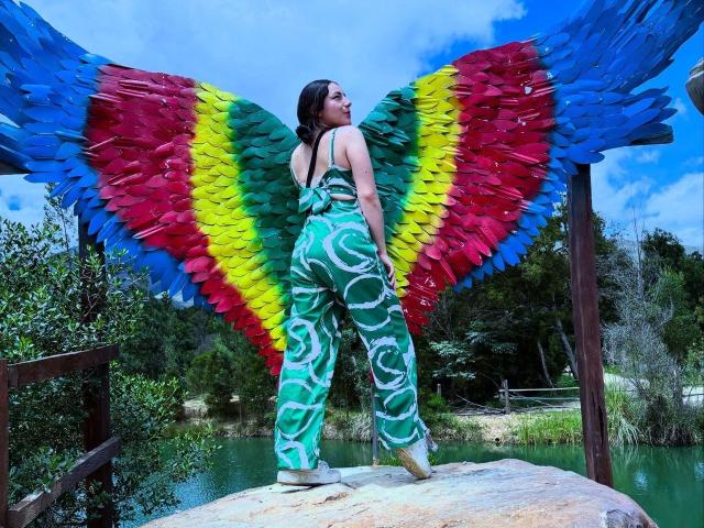 MilaWilde stands on a rock with huge colorful wings, wearing green and white patterned pants, and a black top, looking back with her ass facing the camera.