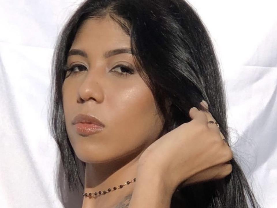 Morenajs has long black hair, tan skin, and glossy lips, with a hand touching her hair. She's wearing a bead necklace and has a tattoo on her shoulder.