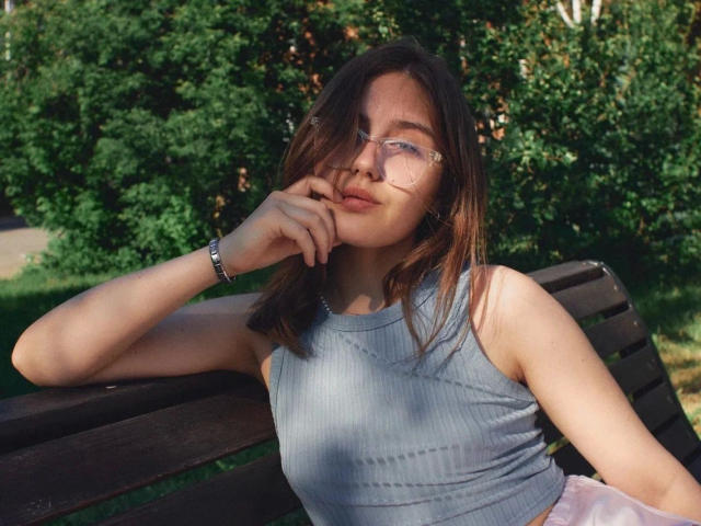 TessaLove, atractiva, con gafas, cabello castaño y piel clara, vestida con una camiseta gris sin mangas, apoyando el mentón en un banco de madera, con una expresión ligeramente aburrida, y con una pulsera en la muñeca..