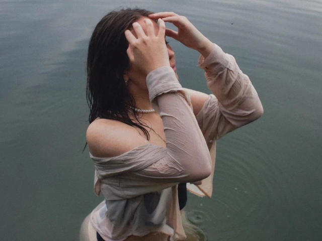 TessaLove, capelli castani bagnati e sensuale, mani che coprono la testa, indossa una camicia beige con una spalla scoperta, una collana, è in piedi in acqua verde, con il seno visibile, la camicia aderente al corpo..