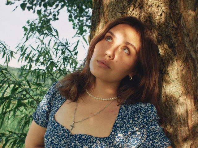 TessaLove-hot with brown hair, blue floral dress, pearl necklace, and small tits, leaning against a tree, looking up with a slightly pouty face, sunlight filtering through.