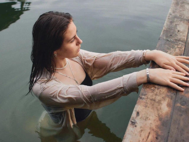 TessaLove – sinnlich, mit nassen, braunen Haaren, trägt ein durchsichtiges Hemd, ein schwarzes Oberteil, eine Perlenkette und ein Armband, lehnt sich an eine Holzleiste und blickt seitlich in grünes Wasser..