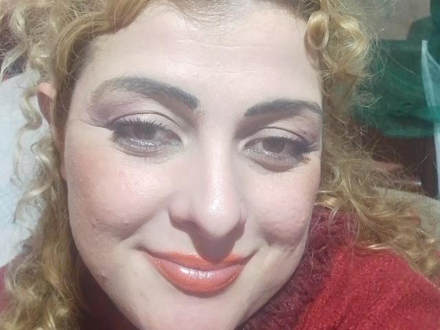 CrislimaJb has curly blonde hair, thick eyebrows, and orange lipstick, smiling with slightly winking eyes. She's wearing a red textured top. Her face is slightly flushed.