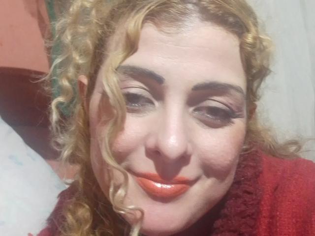 CrislimaJb has curly blonde hair, red lipstick, and thick eyebrows. She's wearing a red textured sweater with a shy smile. Her makeup includes bold eyeliner.