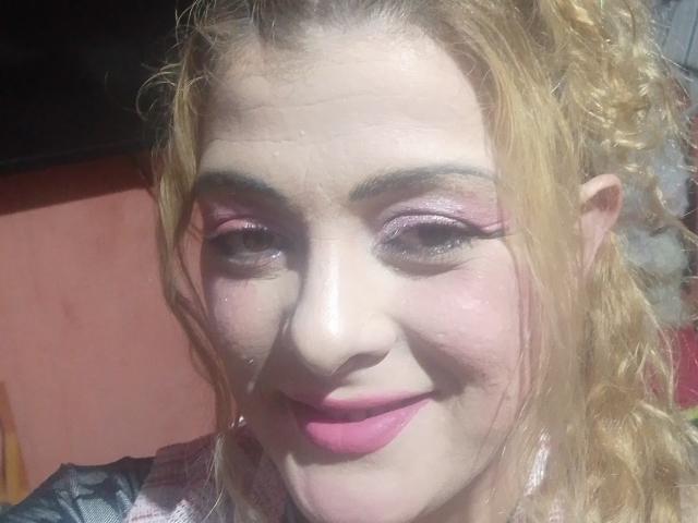 CrislimaJb with curly blonde hair, pink eyeliner, and pink lipstick, smiling at the camera, showing a slightly sweaty face, wearing a plaid shirt.