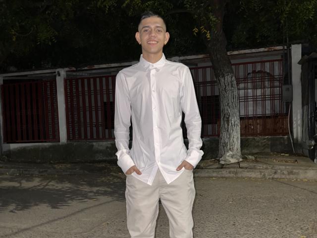 MikeCree stands with hands in white pants pockets, wearing a white button-up shirt, smiling. He's a slim, young Asian dude with short black hair, in a dark outdoor setting.