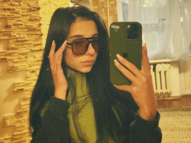 OliviaFoxy with long black hair, wearing sunglasses and a green turtleneck, holding a green iPhone, touching her face, standing in a warmly lit room.