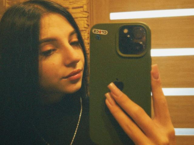 OliviaFoxy with long black hair, holding a green iPhone, taking a selfie. She's looking down at the phone, her finger on the screen, with a subtle smile.
