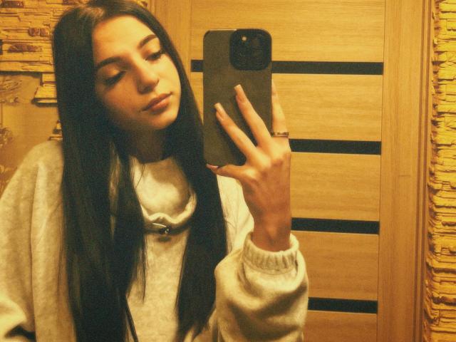 OliviaFoxy with long black hair, holding a phone, wearing a grey sweater, taking a mirror selfie, showing her ring, with a soft, warm-toned, slightly blurry photograph.