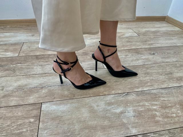 Los pies de Kripkoa, con tacones altos negros brillantes y tiras en el tobillo, mostrando un pequeño tatuaje en el tobillo, vestida con pantalones blancos sobre un suelo de madera..