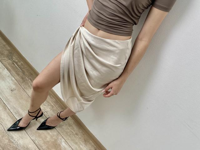 Kripkoa wearing a beige skirt with a thigh-high slit, brown top, black pointed heels, standing on wooden floor, hand on skirt, showing her bare ass and a hint of pussy.
