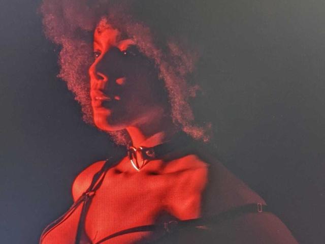 AfroDiva with curly red-lit hair, wearing a black strappy harness and choker, has a strong, defined jawline and visible collarbones. Red light highlights her dark skin.
