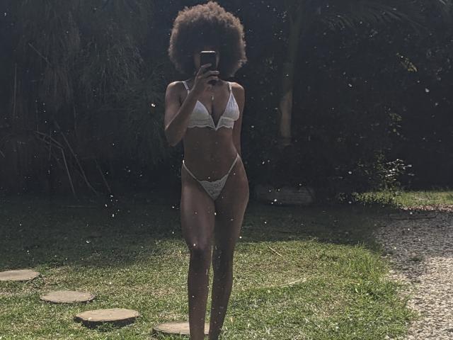 AfroDiva with a curly afro, wearing a white bikini, stands in a grassy yard, holding a phone, showing her small boobs and tiny white bikini bottom.