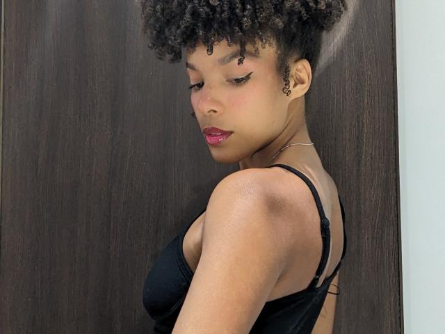 AfroDiva with curly hair, black eyeliner, and red lipstick, wearing a black bra, looking down with her back to the camera, showing her tits and a small tattoo on her cheek.