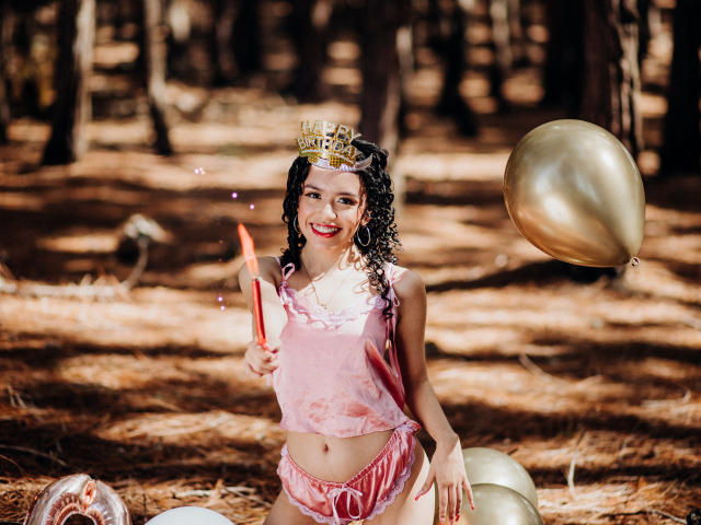 Macaroux with curly black hair, pink velvet top, and matching shorts, holding a sparkler, wearing a gold crown, smiling, with a gold balloon floating near her.