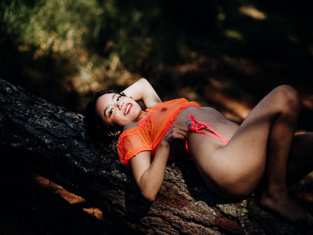 Macaroux is lying on a tree, wearing an orange mesh top and red thong, with her hand on her pussy, looking up with red lips, dark hair, and a smile.