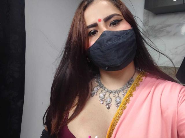 DaliaDubey with brown hair, wearing a black mask, pink outfit, and ornate necklace, has a red dot on her forehead and is showing her tits.