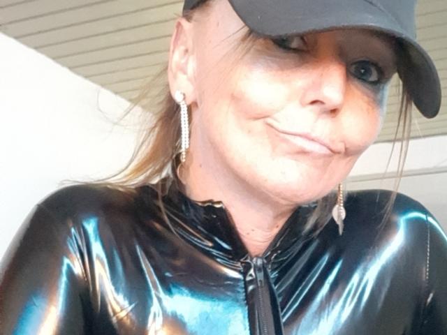 MissFreeBird with shiny black latex top, subtle smile, earrings, and black cap, showing off her puffy lips and freckles. Her tits are covered, but she's looking hot.