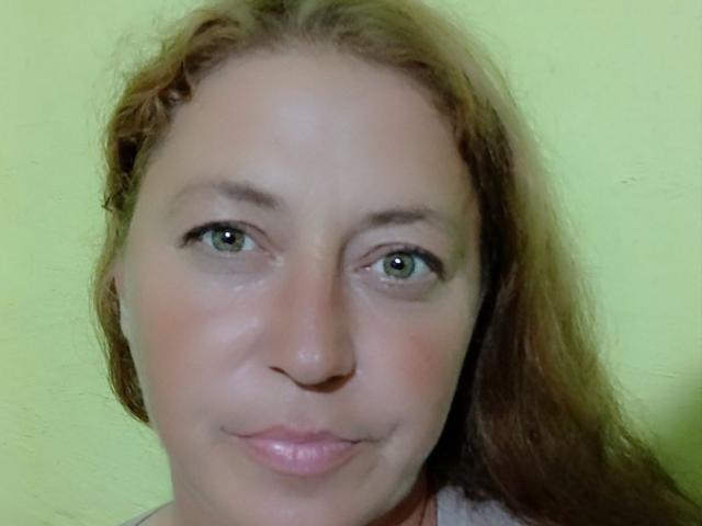 ClaireApple's face with green eyes, light brown wavy hair, slight smile, and pinkish lips. Her skin is fair, and she's wearing a white shirt.