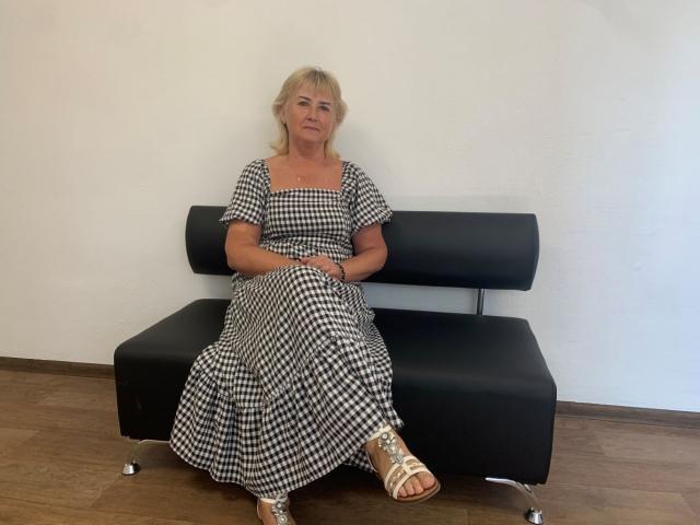 NoraVeda heeft blond haar, een geruit jurkje aan en zit op een zwarte leren bank, waarbij haar billen op de onderste zitting rusten. Ze draagt witte sandalen en zit op een houten vloer..