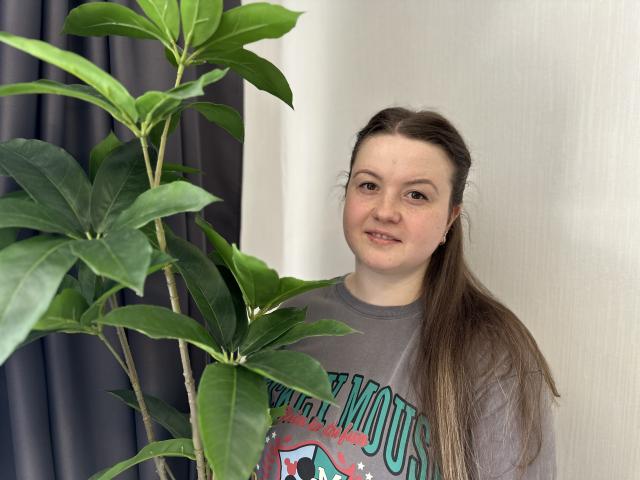 Mavka: Een schattig meisje met een lange bruine paardenstaart, een grijze trui, die naast een grote groene kamerplant staat, zachtjes lachend, bruine ogen, lichte huid, casual en ontspannen..
