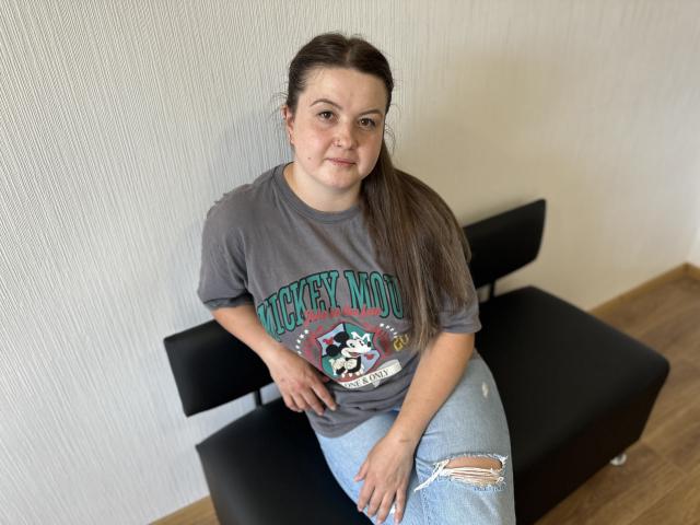 Mavka, ein niedliches, etwas molliges Mädchen mit heller Haut, braunen Haaren im Pferdeschwanz, trägt ein graues T-Shirt mit der Aufschrift "Devil Driver" und zerrissene Jeans. Sie sitzt auf einer schwarzen Bank und lächelt leicht..