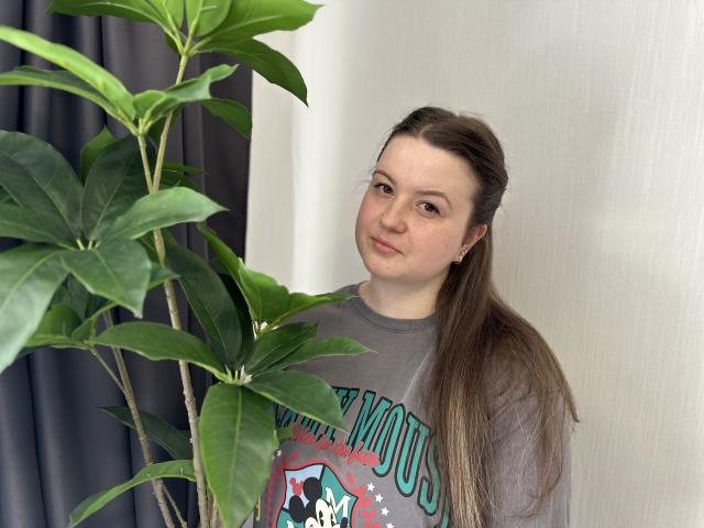 MavkaCute, mit langen braunen Haaren, steht neben einer grünen Pflanze, trägt ein graues T-Shirt mit einem Aufdruck und hat ein leichtes Lächeln auf ihrem leicht rundlichen Gesicht..