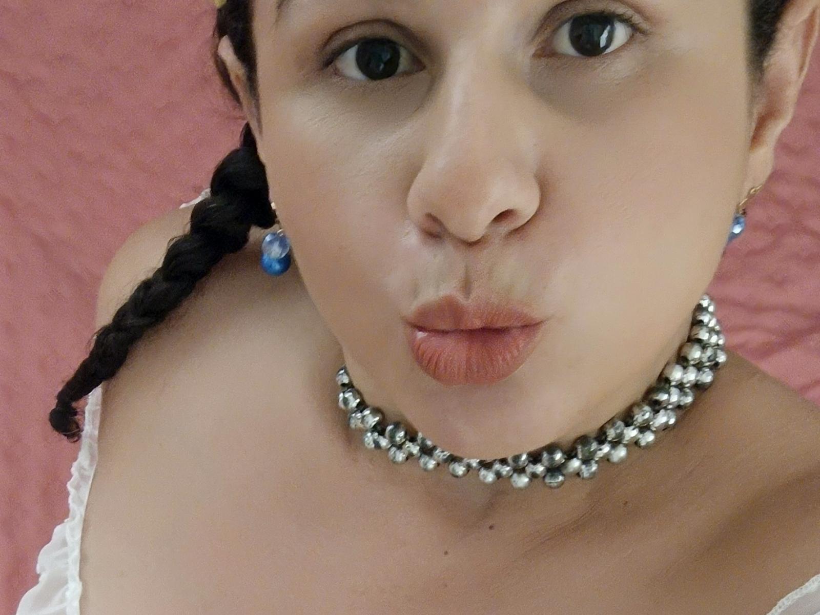 Discordia with pouty lips, wearing a silver bead necklace, white strap, and blue earrings, has a black braid, and pale skin, looking up with blue eyes.