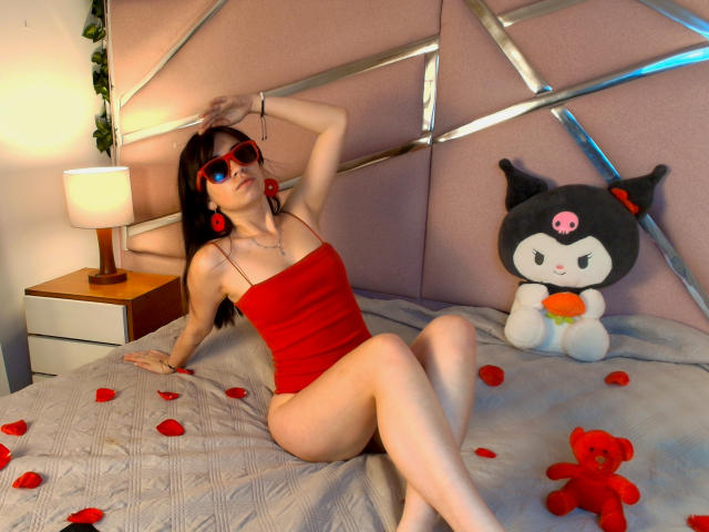 AnyuWu with red sunglasses, red dress, and red earrings, sitting on bed with rose petals, legs crossed, hand on head, toy black cat with pink nose, and red teddy.