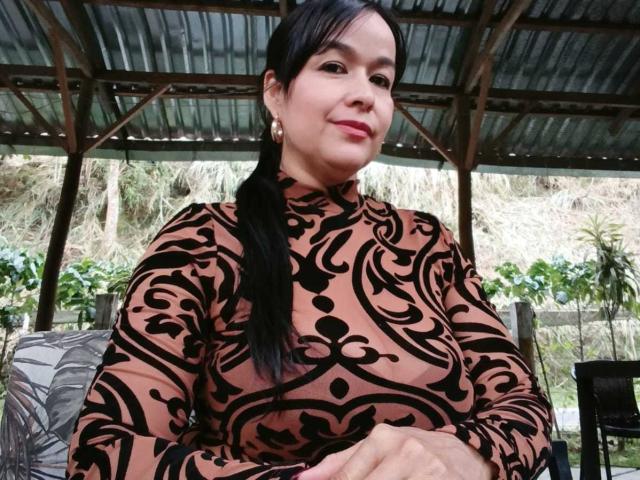 TamaraFoxy with black ponytail, brown and black patterned dress, small tits, smiling, wearing gold earrings, under metal roof, hands on lap, green plants in background.