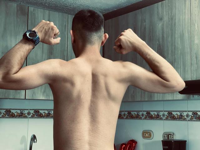 MartinBlos with a muscular back, flexing both arms, wearing a black wristwatch, short dark hair, and a light skin tone, in a kitchen.