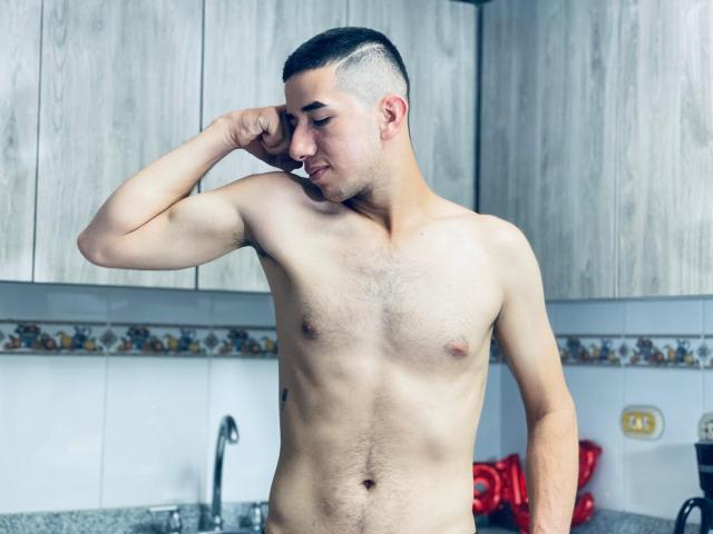 MartinBlos, shirtless with a shaved undercut, flexing his bicep, has a light skin tone, small chest, and a slight smile, standing in a kitchen.
