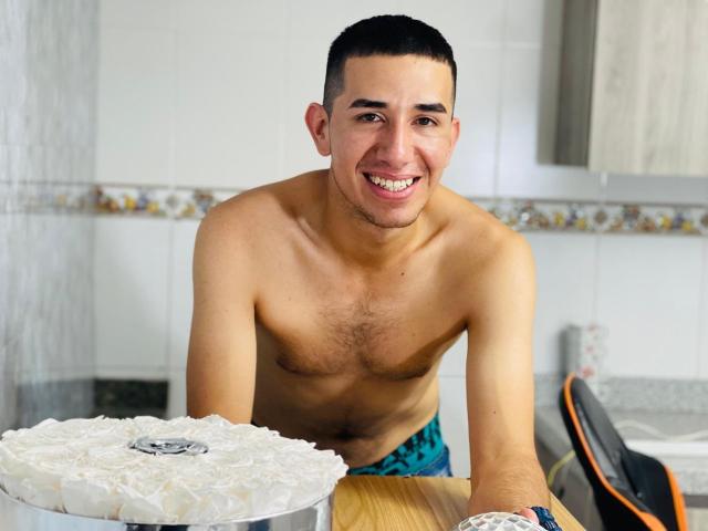 MartinBlos, shirtless with a slight chest hairy, smiling, leaning on a kitchen counter with a frosted cake in front of him, wearing blue and black patterned shorts.