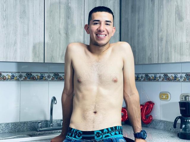 MartinBlos, shirtless, smiling, wearing blue boxer briefs with "CHAMPION" on the waistband, standing in a kitchen, grey wood cabinets, granite countertop, short dark hair, medium build, small chest.