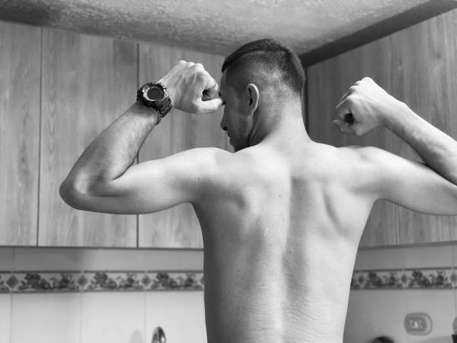 MartinBlos, shirtless, flexing muscles, back to camera, short dark hair, small tattoo on right arm, wearing a watch, in a kitchen, raising fists, grey-scale photo.