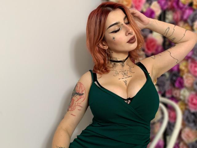 HannahSexy69 with red hair, green dress, big tits, tattoos, nose ring, and makeup, standing against white wall, hand in hair, looking down, floral pattern in background.