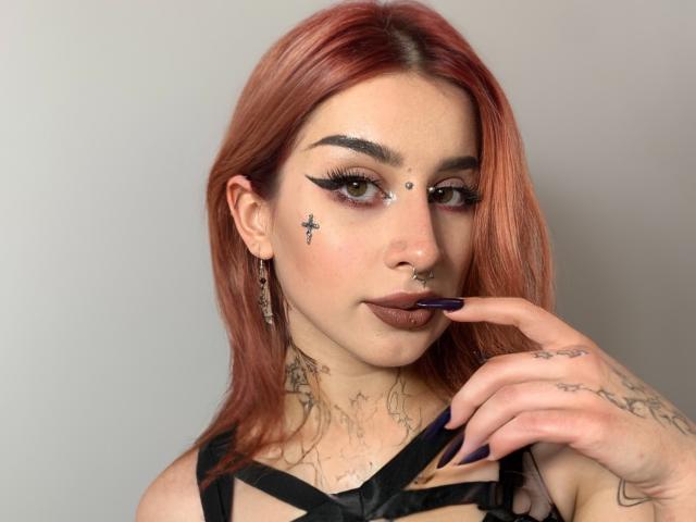 HannahSexy69 with red hair, dark makeup, nose ring, tattoos, black strappy top, purple nails, finger to lips, looking at camera. Boobs visible, tattoos on neck and hand.